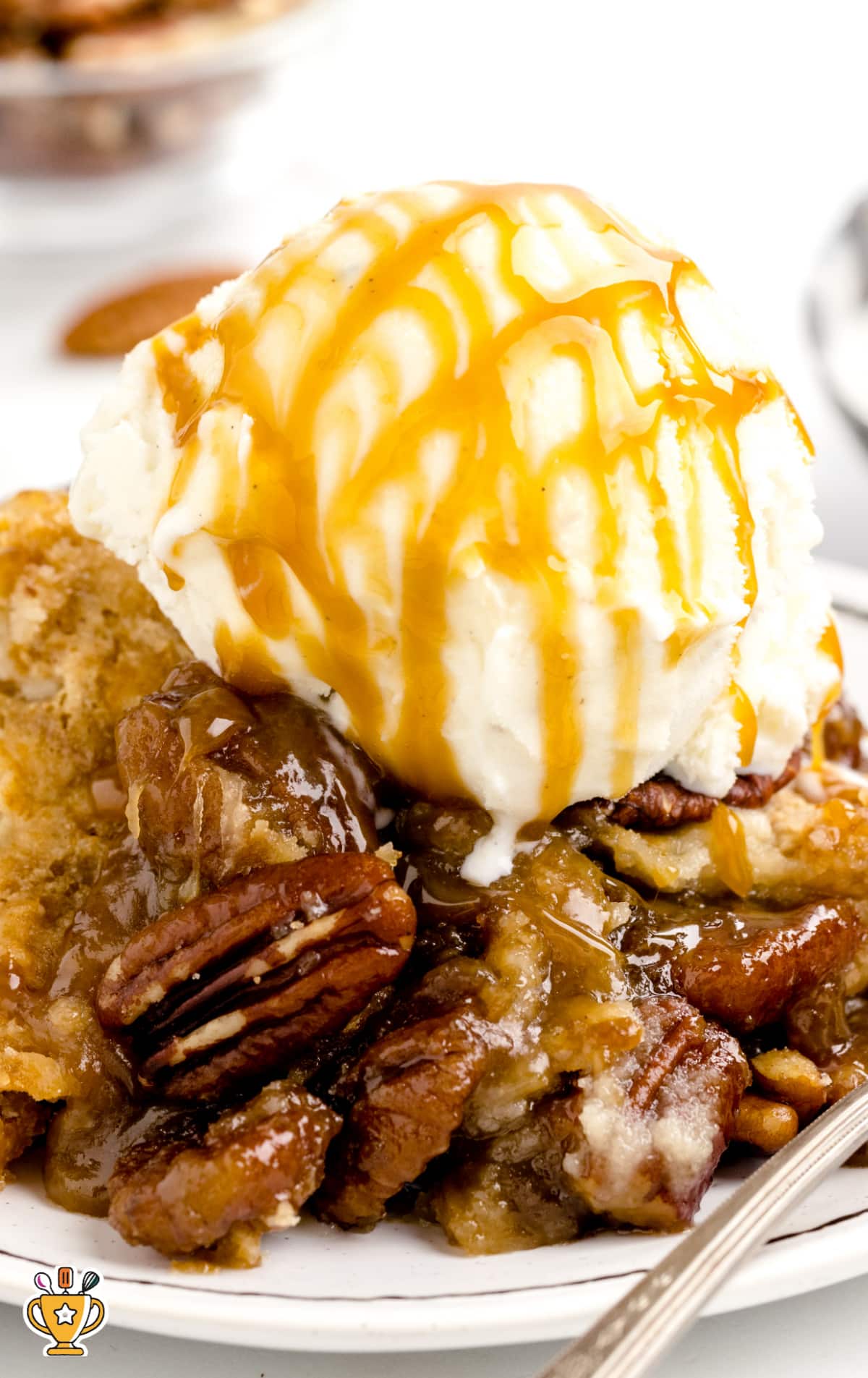 A close-up of a warm serving of pecan dump cake topped with a generous scoop of vanilla ice cream and drizzled with golden caramel sauce, highlighting the gooey texture and glossy pecan topping on a white plate with a spoon.