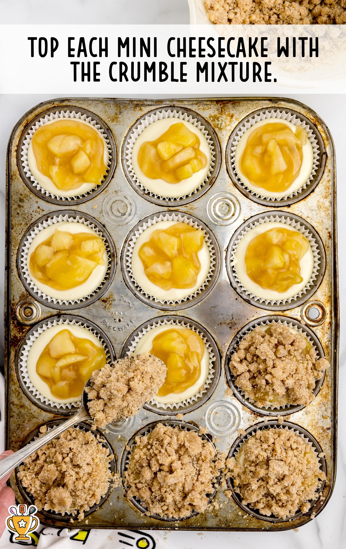 A hand holding a spoon full of crumble mixture adding on top of the mini Cheesecakes