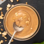 A jar of 3 ingredient satay sauce with a spoon it in, and crushed peanuts on top.