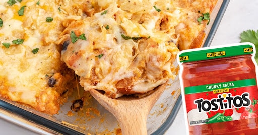 A casserole dish full of chicken taco casserole with a serving spoon, near a jar of salsa.