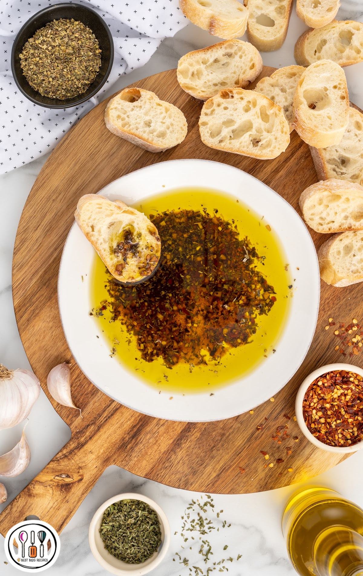 An overhead shot of a serving board with bread slices, a bowl of Olive Oil Bread Dip, and seasoning.