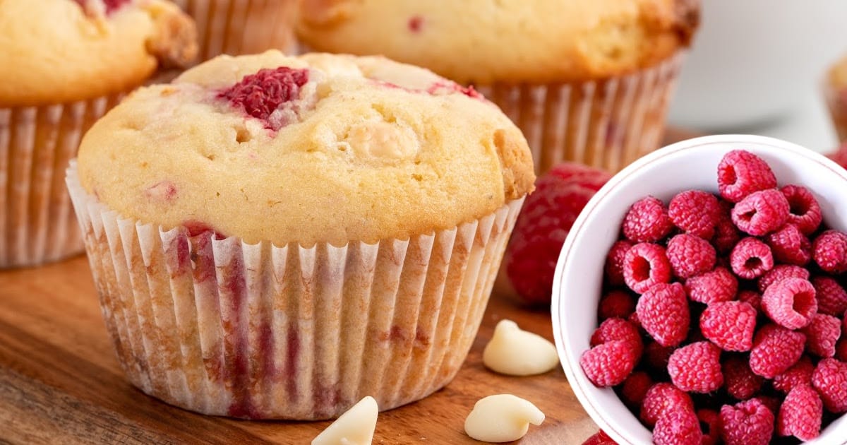 A White Chocolate and Raspberry Muffin on a cutting board beside a white bowl full of fresh raspberries.