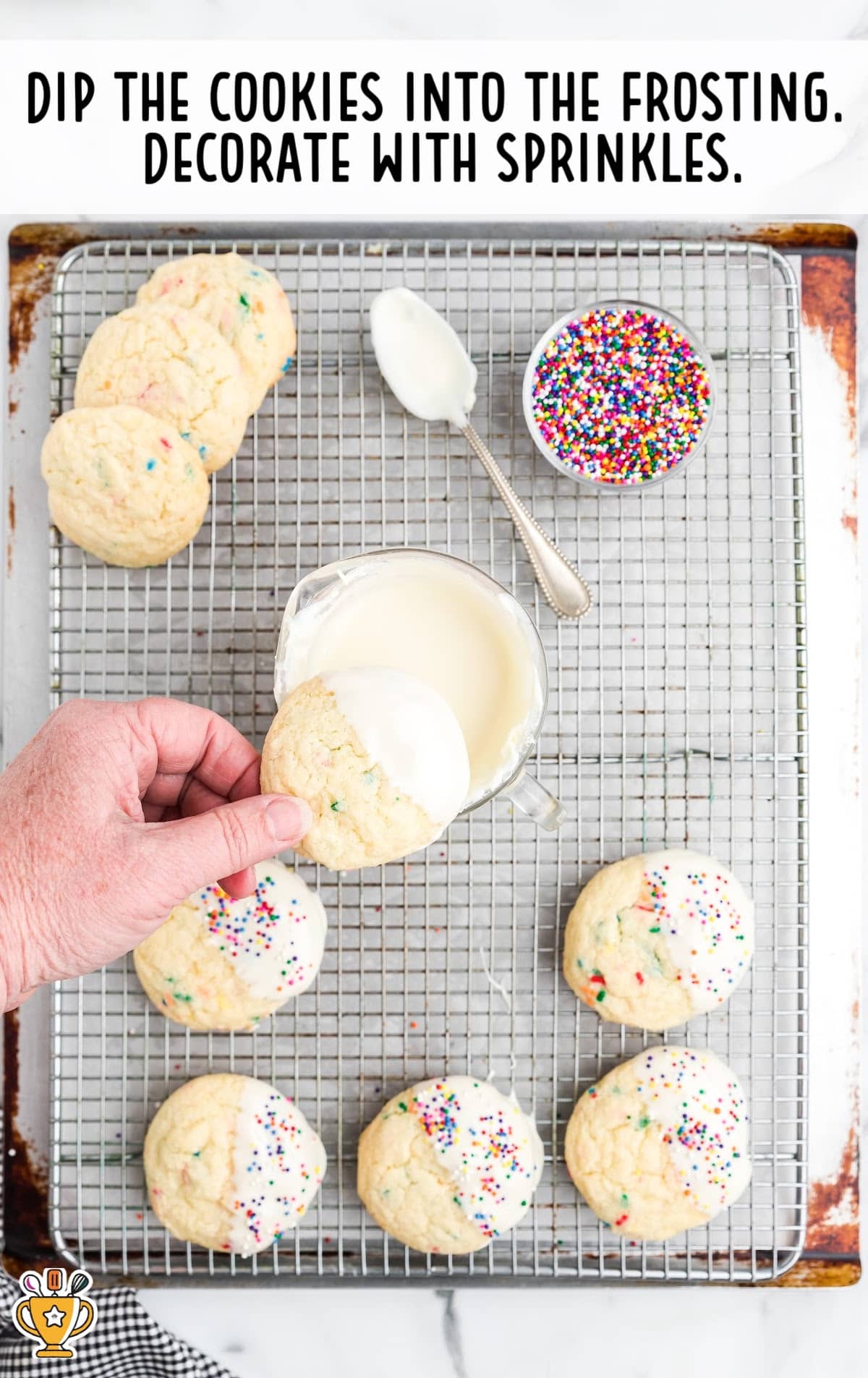A hand dipping cookies into the frosting, with a bowl of sprinkles and a spoon covered with frosting on the side