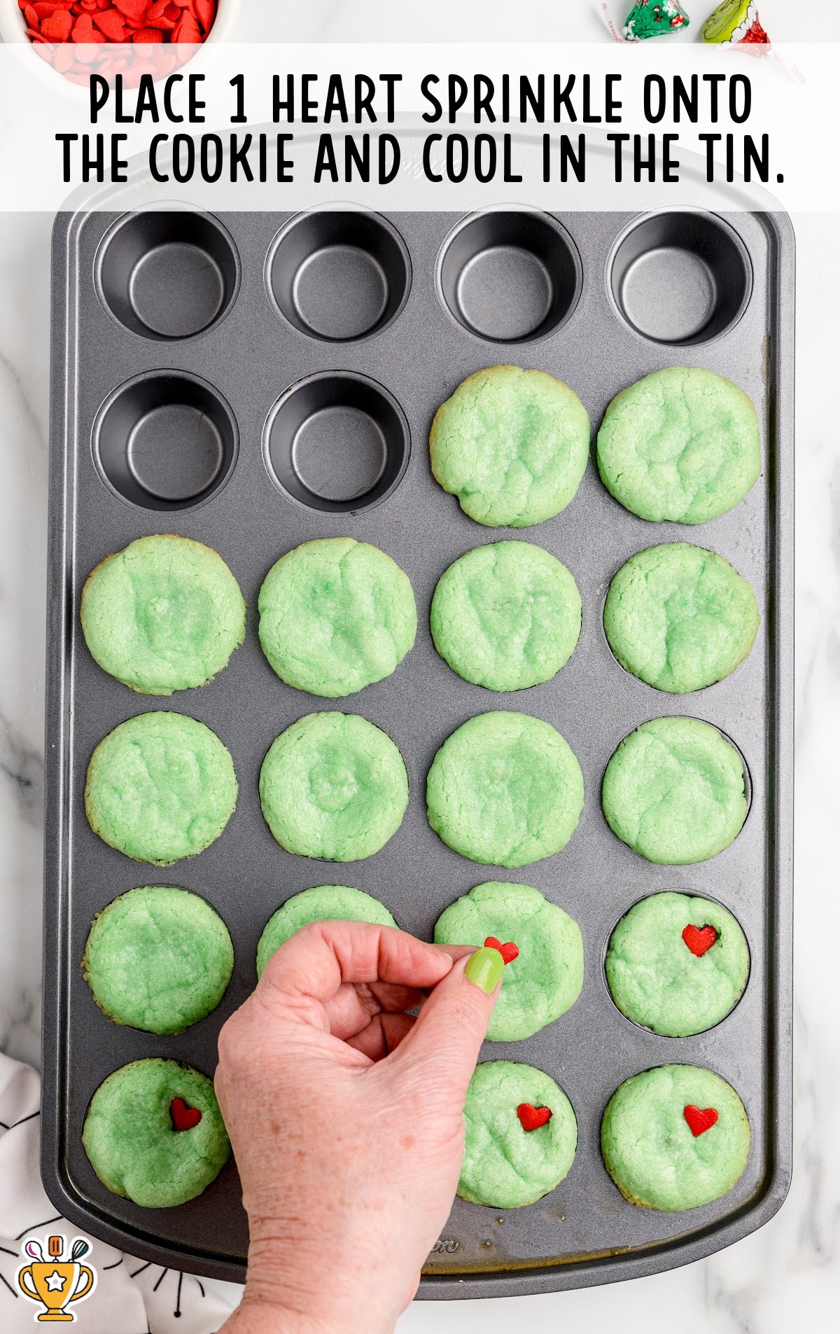 Grinch Cookies in a cupcake pan with a hand placing heart sprinkles on top of the cookies