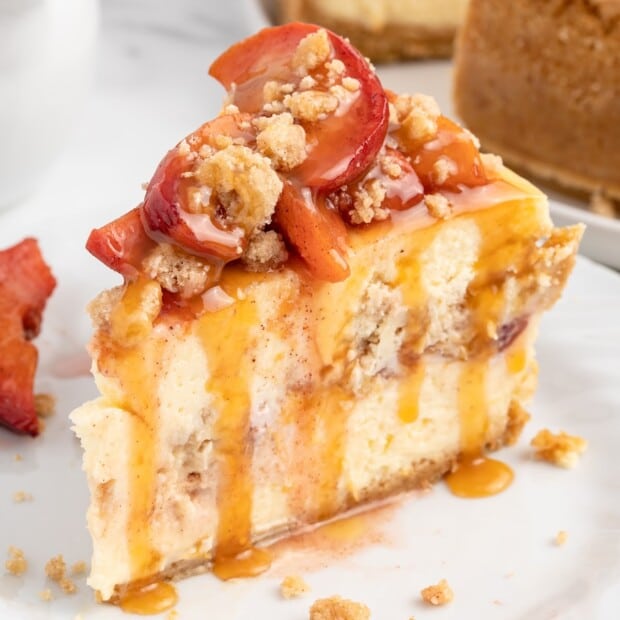 Square photo of a peach cobbler cheesecake slice on a white plate, showing creamy layers with peach filling, caramel drizzle, and a chunky peach crumble topping, with the whole cheesecake blurred in the background.