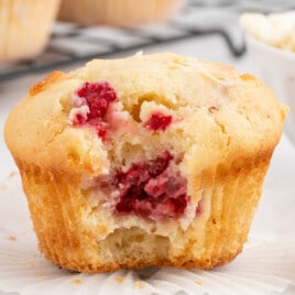 A white plate with a White Chocolate and Raspberry Muffin on it. There's a bite out of it, showing the white chocolate and raspberries inside.