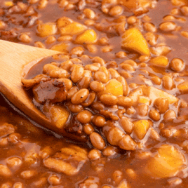 Close-up of apple pie baked beans in a skillet, featuring tender baked beans, chunks of apple, and crispy bacon pieces in a rich, glossy brown sauce, with a wooden spoon scooping out a hearty serving.