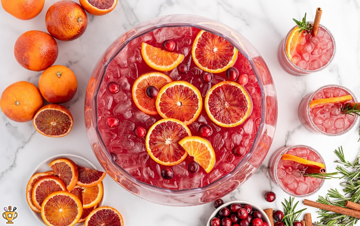 Overhead view of a large bowl of cranberry Christmas cocktail filled with ice, blood orange slices, and cranberries, surrounded by festive garnished glasses.