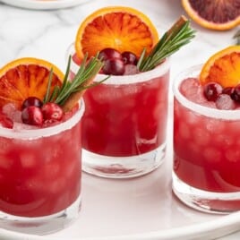 Close-up of a cranberry Christmas cocktail in a rocks glass with crushed ice, garnished with blood orange, fresh cranberries, rosemary, and a cinnamon stick.