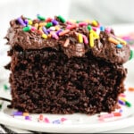 Close-up of a slice of moist chocolate crazy cake topped with rich chocolate frosting and colorful rainbow sprinkles, served on a white plate with a few extra sprinkles scattered around.