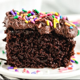 Close-up of a slice of moist chocolate crazy cake topped with rich chocolate frosting and colorful rainbow sprinkles, served on a white plate with a few extra sprinkles scattered around.