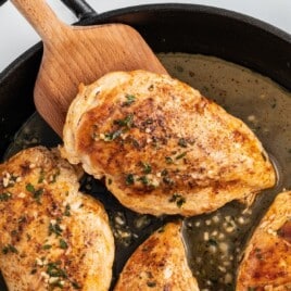 a wooden cooking spoon holding one Pan Seared Chicken piece and others in a skillet