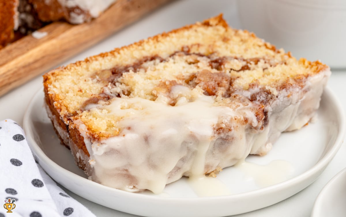 Close-up of a thick slice of apple fritter bread on a white plate, showing a soft vanilla crumb layered with cinnamon-sugar apple swirls and finished with a generous drizzle of white glaze that pools slightly on the plate, styled with a neutral kitchen background and a folded polka-dot cloth nearby.