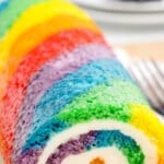 Close-up of a colorful rainbow roll cake with vibrant, spiraled layers of red, orange, yellow, green, blue, and purple sponge cake, filled with creamy white buttercream, sliced and served on a plate.