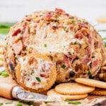 A cheese ball is sliced with a knife and a cracker is covered in the yummy cheese.
