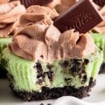 Mini Mint Cheesecake with a crushed Mint Oreo crust, creamy green mint cheesecake filling, topped with swirls of chocolate whipped topping and a square of Andes mint chocolate, with a bite taken to reveal the texture.