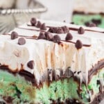 Slice of Mint Chocolate Chip Ice Cream Cake with layers of crushed chocolate cookie crust, green mint chocolate chip ice cream, whipped topping, chocolate drizzle, and mini chocolate chips, served chilled with Cool Whip branding shown.