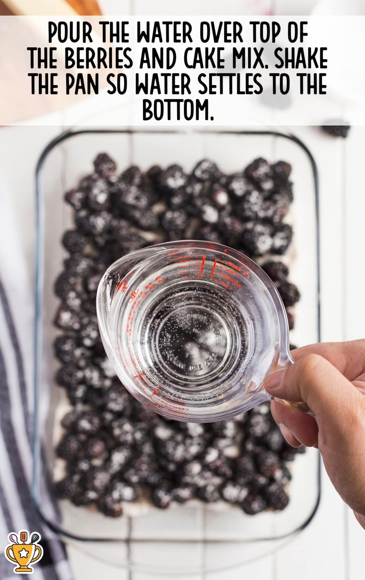 Hand pouring water over cake mix and berries in a baking dish to help it settle