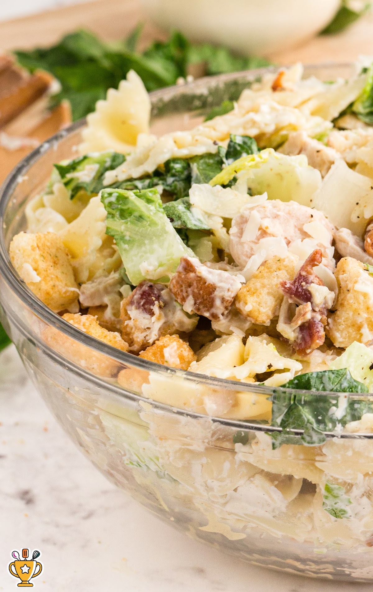 Side view of Caesar pasta salad in a clear bowl showing crisp lettuce, chicken chunks, croutons, bacon, and farfalle pasta.