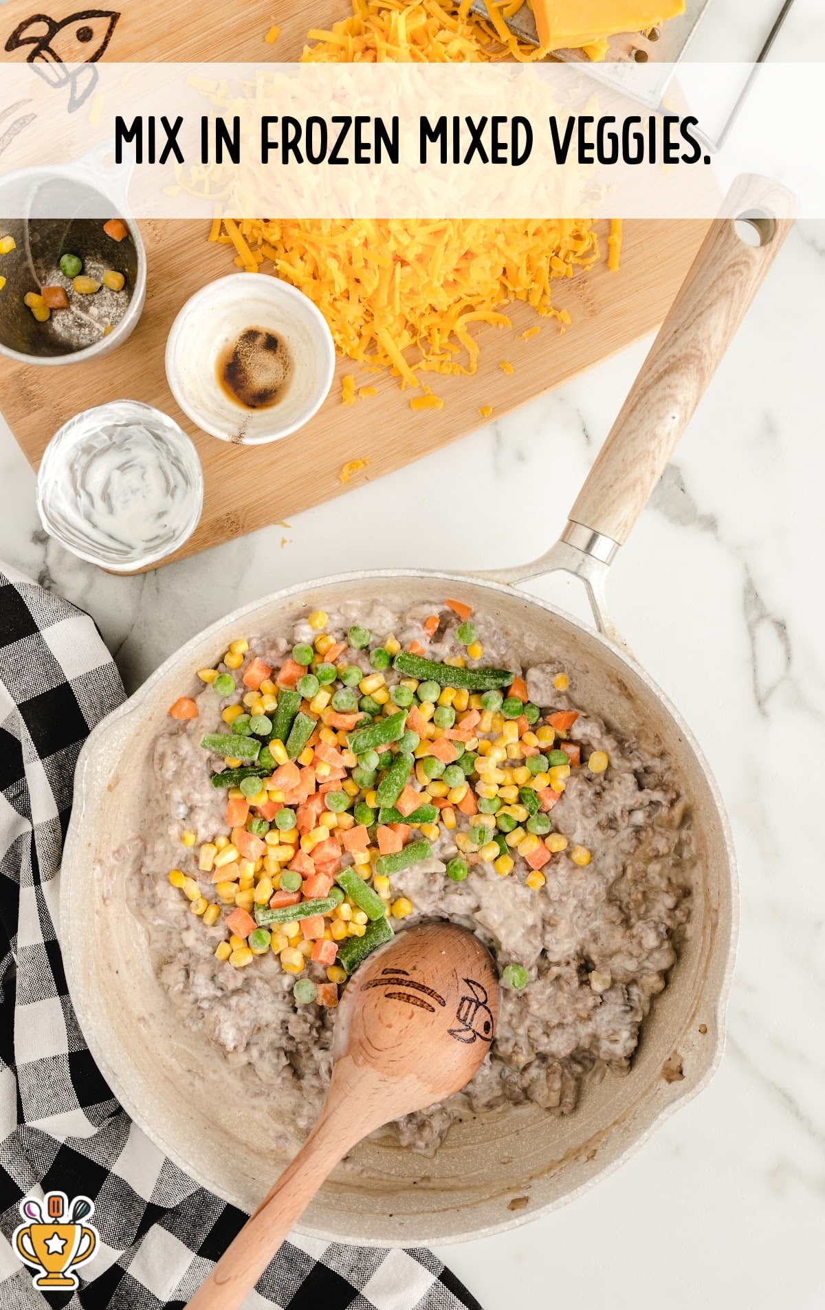 Frozen mixed veggies added on top of creamy beef mixture in skillet. Wooden spoon and cheese board in background.