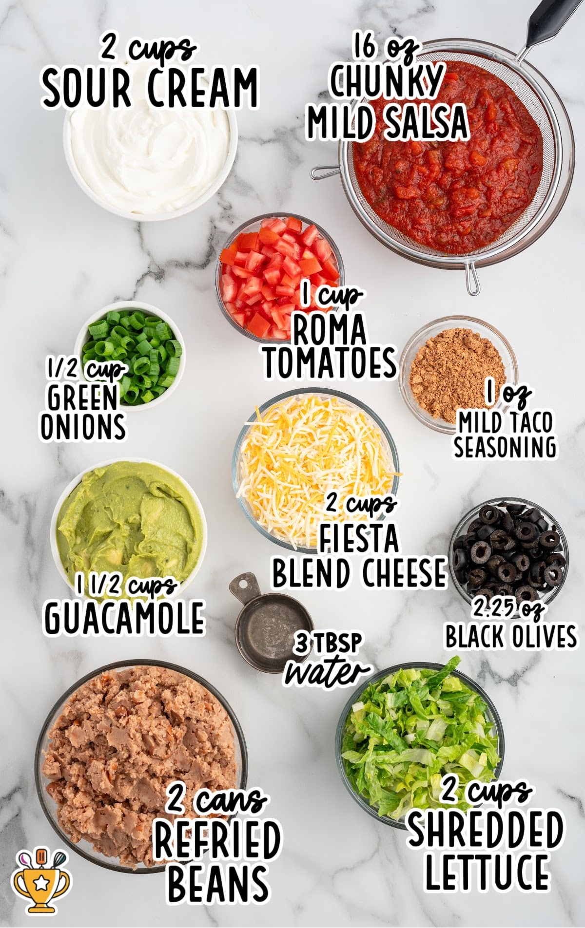 Flat lay of all ingredients for 7 layer dip, each labeled, including sour cream, salsa, guacamole, beans, tomatoes, cheese, olives, lettuce, and seasonings.