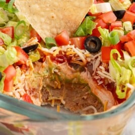 Square close-up of a layered taco dip in a glass baking dish, highlighting colorful layers of refried beans, guacamole, seasoned sour cream, salsa, cheese, lettuce, and tomato.