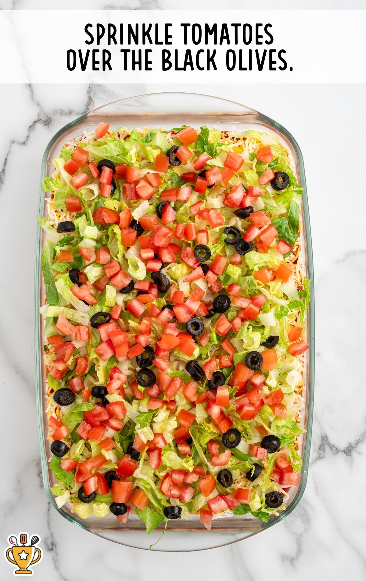 Diced tomatoes are sprinkled over the black olives and lettuce in the glass baking dish, adding vibrant red color to the layered dip.
