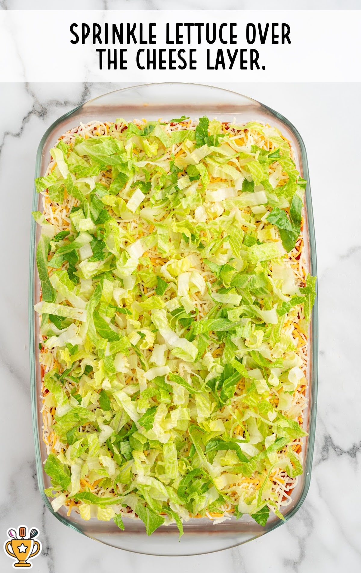 Top-down image of shredded lettuce scattered across the cheese layer in a glass dish. Text overlay says "Sprinkle lettuce over the cheese layer."