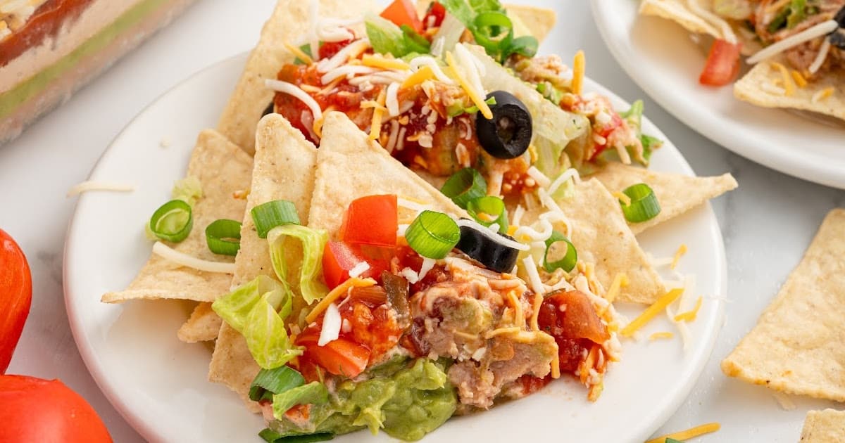 Tortilla chips on a white plate topped with 7 layer dip, featuring bright toppings like tomatoes, olives, lettuce, and cheese.