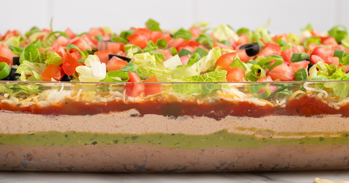 Straight-on shot of a full glass dish of 7 layer dip, emphasizing clean, colorful layers from beans to toppings.