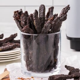 Glass cup filled with air fryer teriyaki beef jerky sticks on a parchment-lined board with spices nearby.