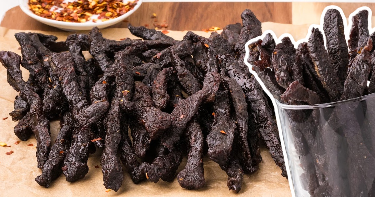 Pile of beef jerky on brown parchment with another bunch of jerky in a glass cup beside it.