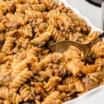 Close-up of a white casserole dish filled with hearty Amish casserole made with rotini pasta, ground beef, and green peppers, with a serving spoon scooping from the pan. Text overlay says &ldquo;Amish Casserole &ndash; Perfect For The Family!&rdquo;