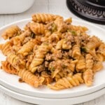 Plate of Amish casserole showing spiraled rotini pasta with tomato-based beef sauce and green peppers, styled on a white wooden background for a recipe card layout.