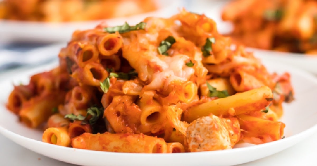 Close-up of a plate of cheesy baked ziti with chunks of chicken and fresh basil, with a small inset image of the same pasta in the lower left corner.