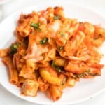 Straight-on view of a white plate filled with baked ziti, chicken chunks, tomato sauce, melted cheese, and sprinkled basil.