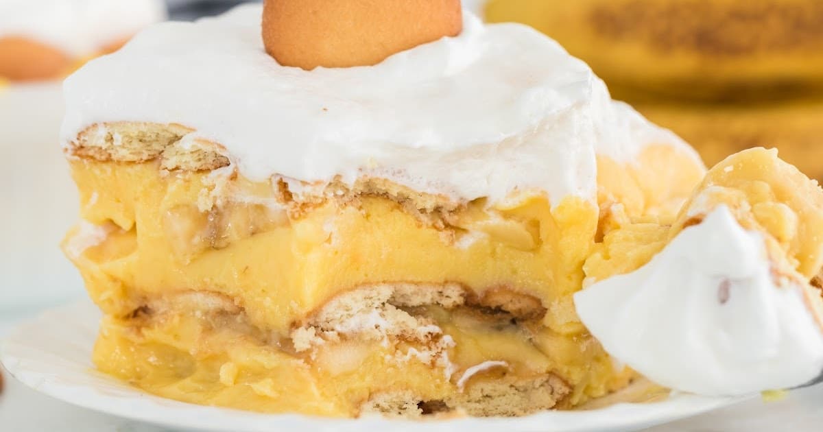 Close-up slice of banana pudding on a white plate with a dollop of whipped topping and a vanilla wafer on top, with a spoonful being scooped.