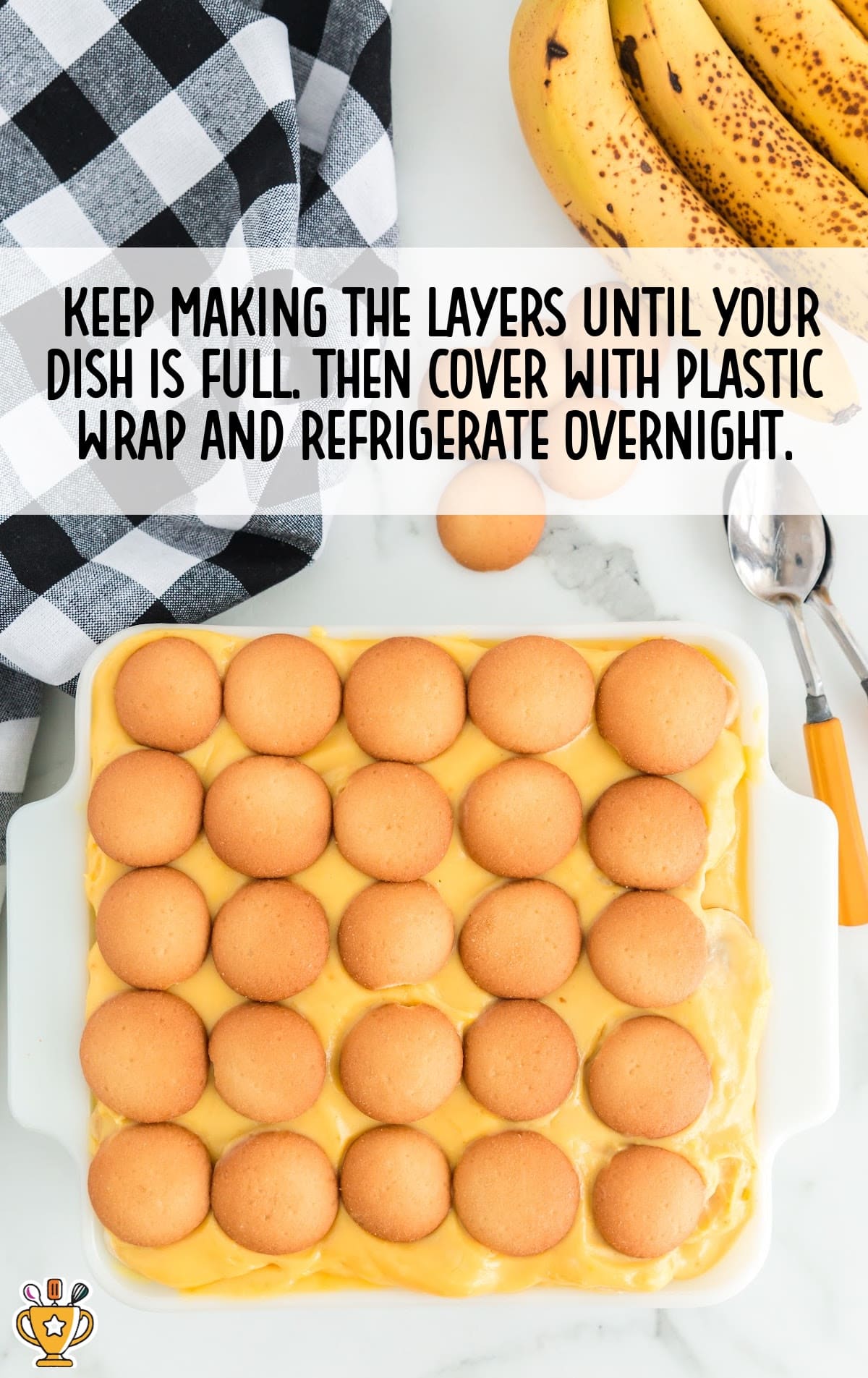 Overhead view of banana pudding layered with vanilla wafers in a white baking dish, surrounded by ripe bananas and a black-and-white kitchen towel.
