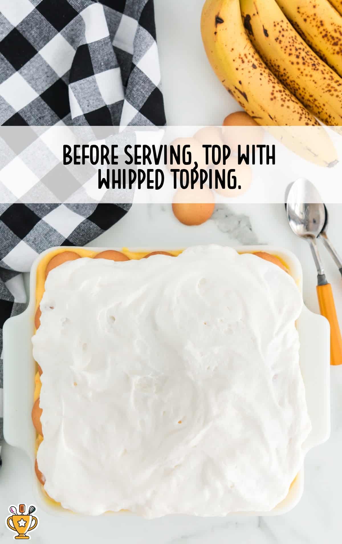 Top-down view of banana pudding topped with whipped topping in a white dish, with bananas and cookies arranged nearby on a white countertop.
