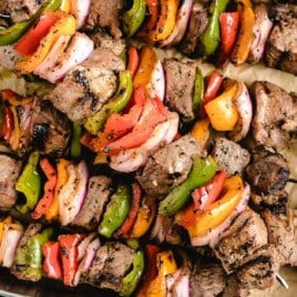 Grilled beef kabobs in a metal tray with parchment paper, filled with skewers of seasoned steak and colorful veggies.