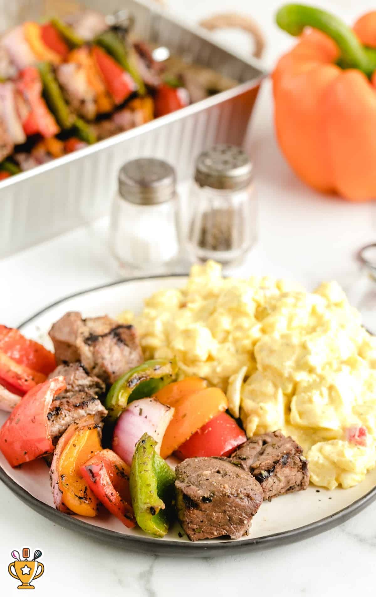 Plate of beef kabobs with creamy potato salad, served next to a tray of skewers and colorful bell peppers.