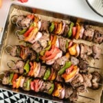 Metal tray filled with grilled beef kabobs, each skewer stacked with beef cubes, colorful peppers, and red onion slices.
