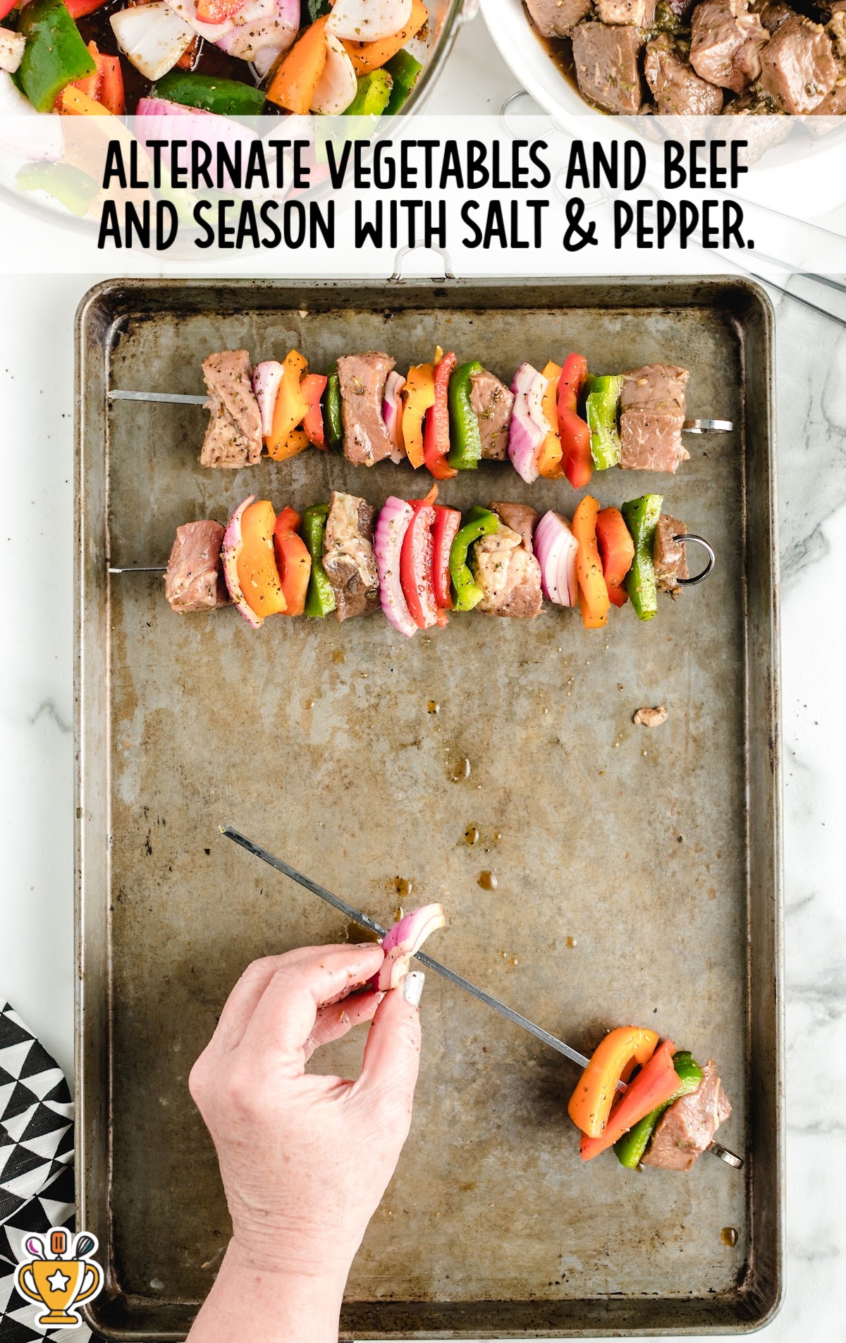 Hand skewering marinated beef and vegetables on a metal skewer over a baking sheet, prepping kabobs for grilling.