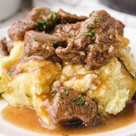 Plate of mashed potatoes topped with tender beef tips and brown gravy, sprinkled with parsley.