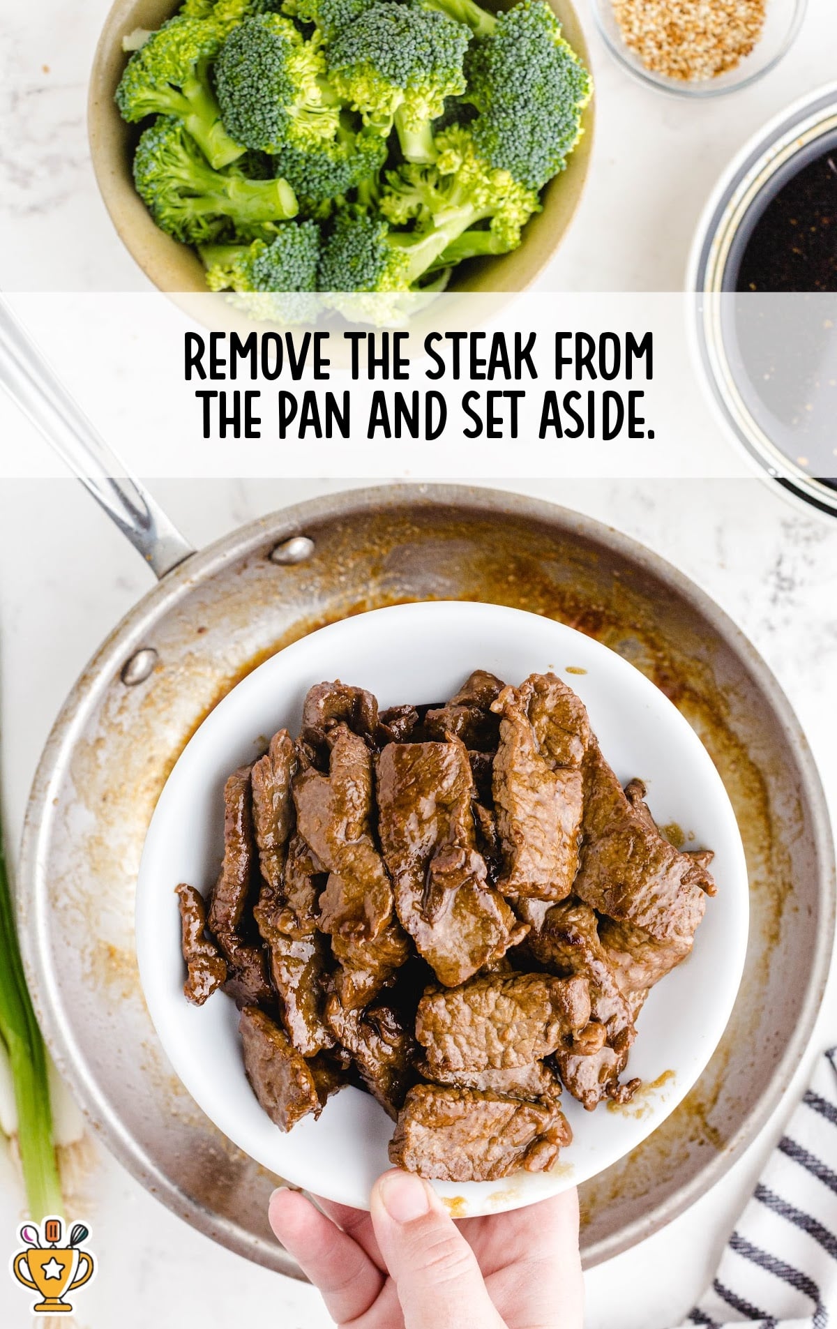 Cooked sliced steak in a white bowl held over a skillet, with fresh broccoli and sauce ingredients in the background.