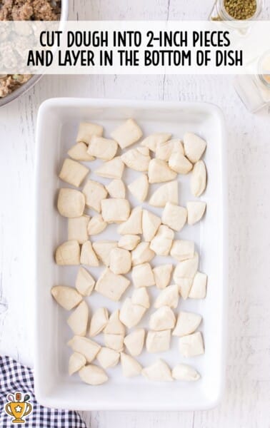 Raw biscuit dough pieces cut into 2-inch chunks and arranged in the bottom of a white baking dish, ready for layering.