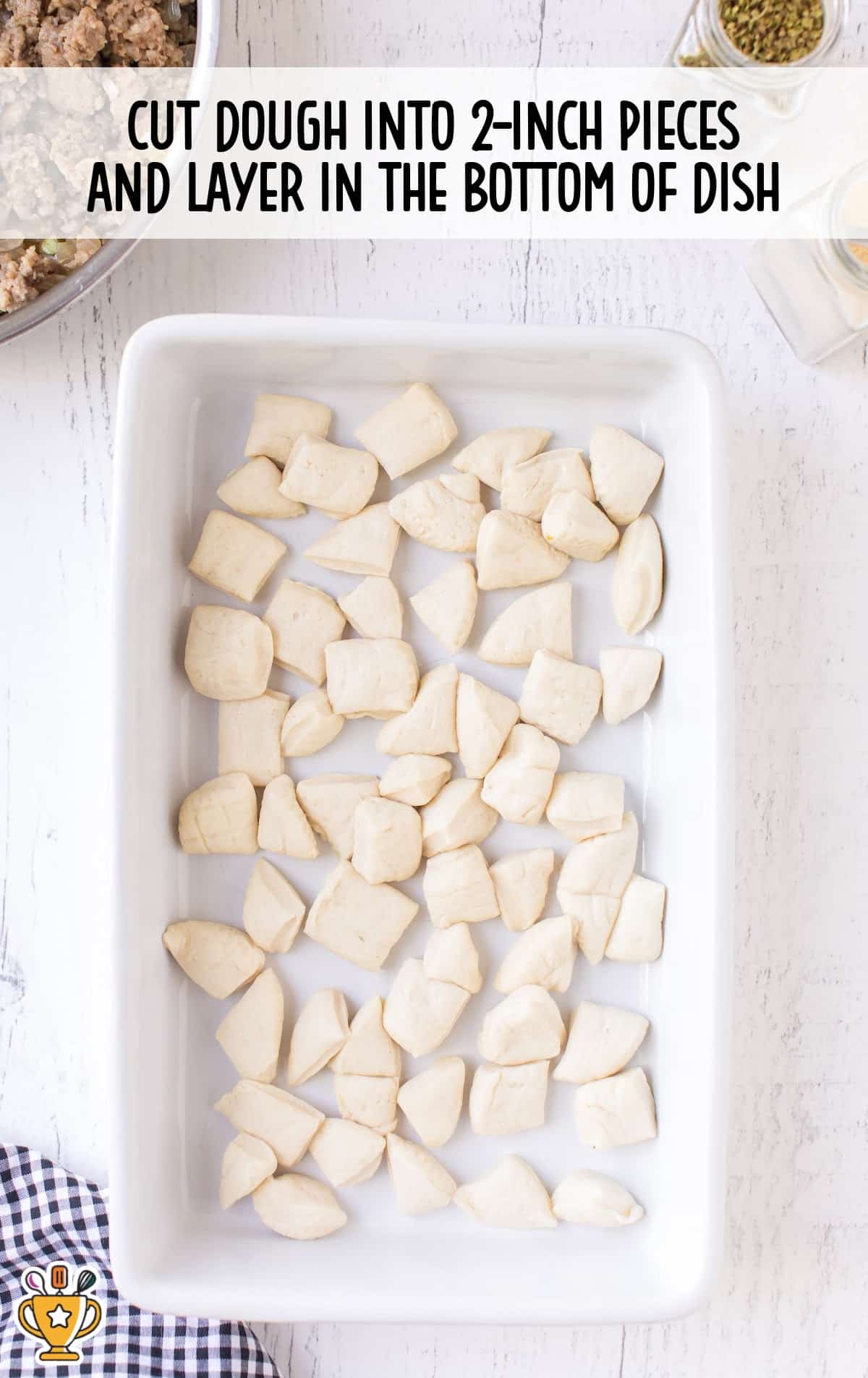 Raw biscuit dough pieces cut into 2-inch chunks and arranged in the bottom of a white baking dish, ready for layering.