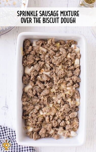 Browned sausage and onion mixture evenly spread on top of biscuit dough in a white casserole dish.