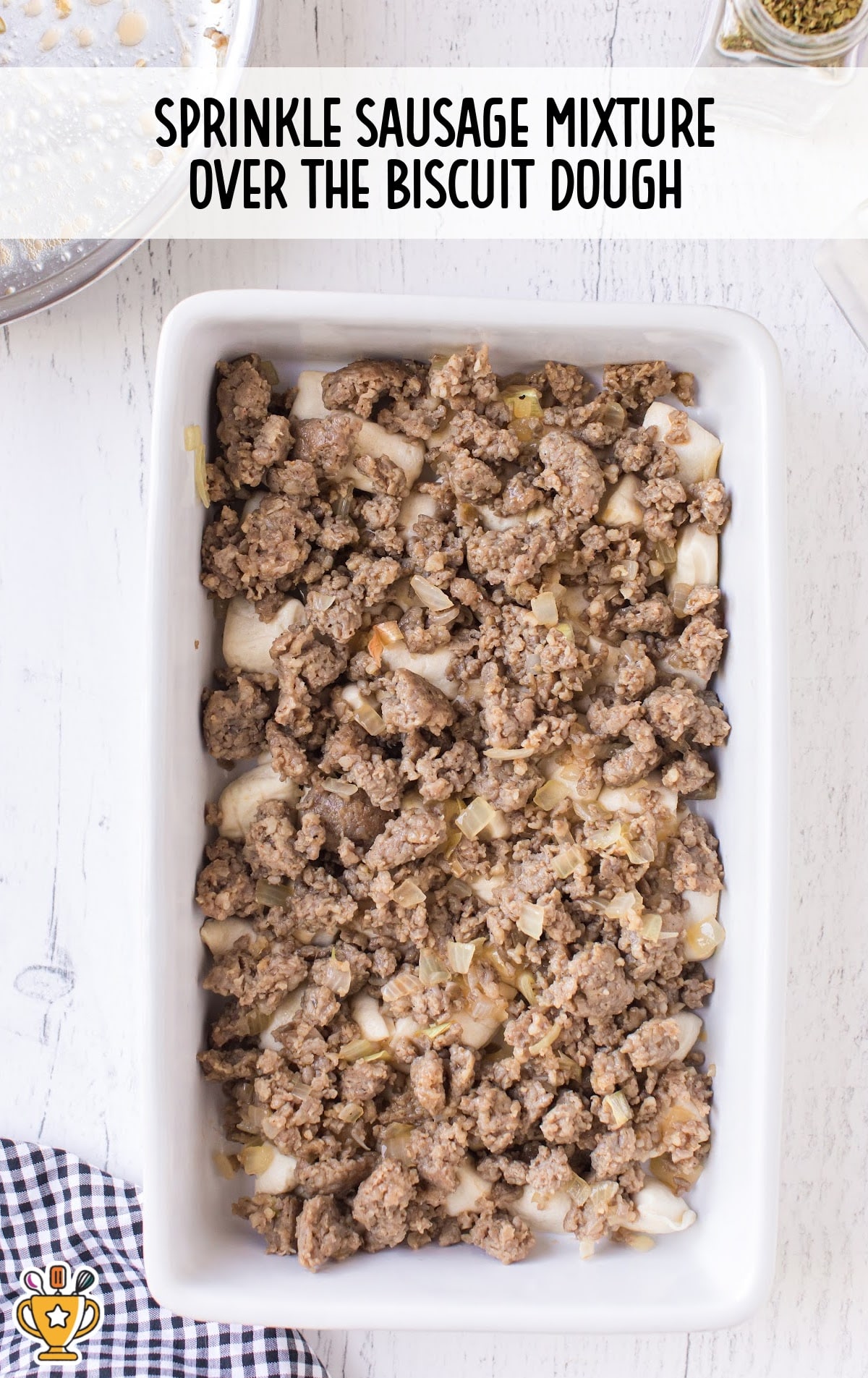 Browned sausage and onion mixture evenly spread on top of biscuit dough in a white casserole dish.