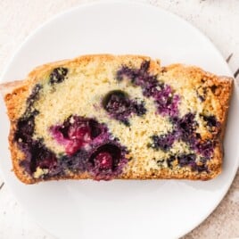 Single slice of homemade blueberry bread on a white plate showing golden crust and juicy baked blueberries inside